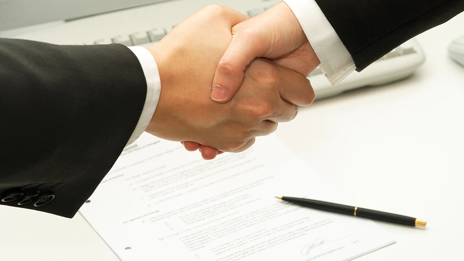 Closeup of the arms of two businessmen shaking hands, with a pen and signed document on a table below them.