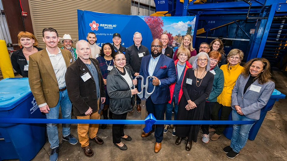 group of people smiling, holding large scissors in front of blue ribbon